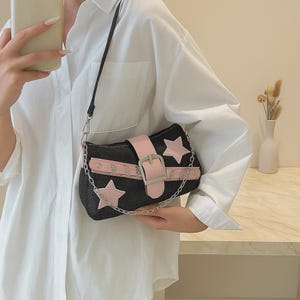 Peut inclure: Sac à bandoulière en jean noir avec des étoiles roses et une sangle rose avec une boucle argentée. Le sac est doté d'une bandoulière en chaîne argentée. Le sac est tenu par une personne portant une chemise blanche.