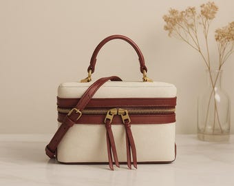 May include: A rectangular cream-colored handbag with a brown leather handle and shoulder strap. The bag features a brown leather trim, gold zippers, and matching zipper pulls. The bag is sitting on a white surface.
