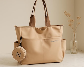 May include: A beige tote bag with brown handles and a small, round coin purse attached. The tote has multiple pockets and the text "Mulati Cool" printed on the front. The coin purse has the letter "N" printed on it.