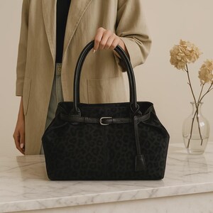 May include: Black tote bag with a leopard print pattern. The bag has two rounded handles and a belt detail with a silver-colored buckle. A small leather tag hangs from the belt. The bag is held in front of a person wearing a tan blazer.