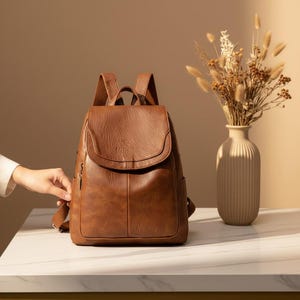 Può includere: Uno zaino in pelle marrone con chiusura a patta e spallacci regolabili. Lo zaino ha una tasca frontale e tasche laterali. La borsa è su una superficie bianca accanto a un vaso con fiori secchi.