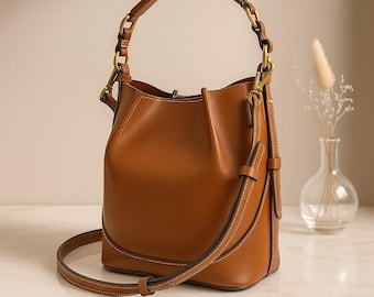 May include: A brown leather bucket bag with a top handle and a detachable shoulder strap. The bag features gold-colored hardware and white stitching details. A small vase with dried flowers is in the background.
