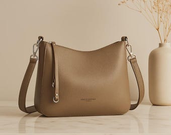 May include: A tan leather shoulder bag with a curved top and adjustable strap. The bag features silver-tone hardware and a small, dangling strap detail. The words "VOLES LEATHER PARIS" are printed on the front of the bag.