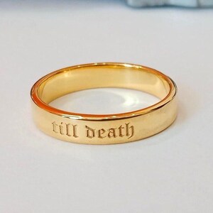 May include: A gold ring with the inscription "till death" engraved in a gothic font. The ring has a smooth, shiny surface and a simple band design. The ring is likely made of gold or a gold-coloured metal.