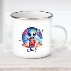 Puede incluir: Taza de esmalte blanco con un extraterrestre de dibujos animados con un traje espacial naranja, saludando. El extraterrestre tiene grandes ojos negros y una cabeza azul y verde. El nombre "Elias" está escrito en guión azul.