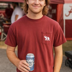 May include: A maroon short-sleeve t-shirt with a white elephant logo and the words "Roll Tide" on the pocket. The person is holding a blue and silver can of beer.