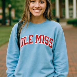 以下が含まれることがあります： 赤と白の文字で「OLE MISS」と書かれた、水色のスウェットシャツ。デニムのショートパンツと合わせています。背景には木々や建物があり、屋外での撮影であることを示唆しています。