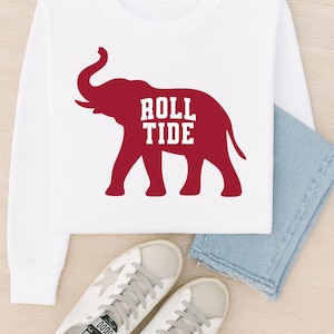 May include: White long-sleeved sweatshirt with a maroon elephant graphic and the text "ROLL TIDE". The sweatshirt is styled with light blue jeans and white trainers with grey laces.