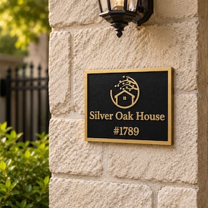 May include: A rectangular house sign with a black background and a gold-colored frame. The sign features a gold-colored graphic of a house outline with a tree above it, followed by the text Silver Oak House and the number #1789 in gold lettering.