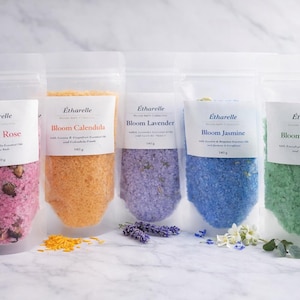 Puede incluir: Cinco bolsas transparentes de sales de baño etiquetadas como "Bloom Rose", "Bloom Calendula", "Bloom Lavender", "Bloom Jasmine" y "Bloom Rosemary". Cada bolsa contiene 140g de sales coloridas y botánicos secos. La marca es "Étharelle".