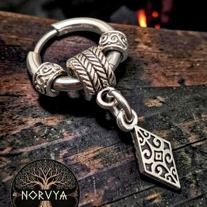 Può includere: Un orecchino a cerchio color argento con due piccole perline cilindriche con volute in rilievo e una perlina centrale più grande con motivo a spina di pesce. Un ciondolo a forma di rombo con un intricato design in filigrana pende dal cerchio.