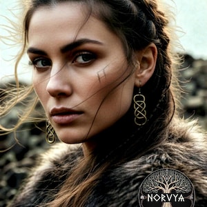 Può includere: Ritratto di una persona con capelli intrecciati, che indossa orecchini d'oro e un indumento simile a pelliccia. Un piccolo tatuaggio è visibile sul viso della persona. L'immagine include il logo "NORVYA" con un disegno ad albero.