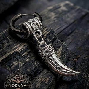 Może przedstawiać: Kolczyk ze srebrzystego metalu z czarną obręczą i wiszącą, zakrzywioną zawieszką w kształcie kła z wygrawerowanymi wzorami. Zawieszka jest przymocowana do obręczy za pomocą ozdobnego srebrnego koralika i małego metalowego kółeczka.