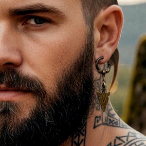 Könnte beinhalten: Nahaufnahme eines Mannes mit Vollbart und einem schwarzen Ohrring mit einem bronzefarbenen Pfeilanhänger. Der Mann hat ein Hals-Tattoo und das Norvya-Logo. Der Ohrring ist im Creolen-Stil mit einem dekorativen Anhänger.