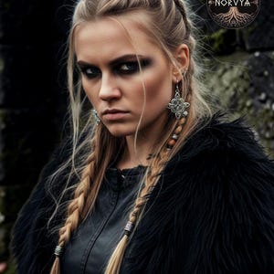 Può includere: Una donna con trecce bionde e trucco scuro, indossa un cappotto di finta pelliccia nero e una giacca di pelle. Indossa anche orecchini d'argento e perline per capelli. Il logo Norvya è visibile.