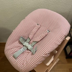 May include: A high chair with a pink and white checkered seat cover and gray safety straps. The wooden frame is a light pink color. The high chair is designed for infants and toddlers.