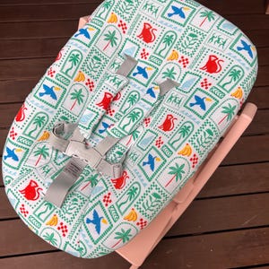 May include: A baby bouncer with a light pink frame and a white fabric cover featuring a repeating pattern of red monkeys, blue toucans, yellow bananas, and green palm trees. The seat includes a gray multi-point harness system with shoulder pads.