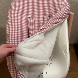 May include: A baby high chair with a removable, padded seat cover. The cover features a red and white gingham pattern on the exterior and a soft white interior. The high chair is white and has a light wood-colored base.