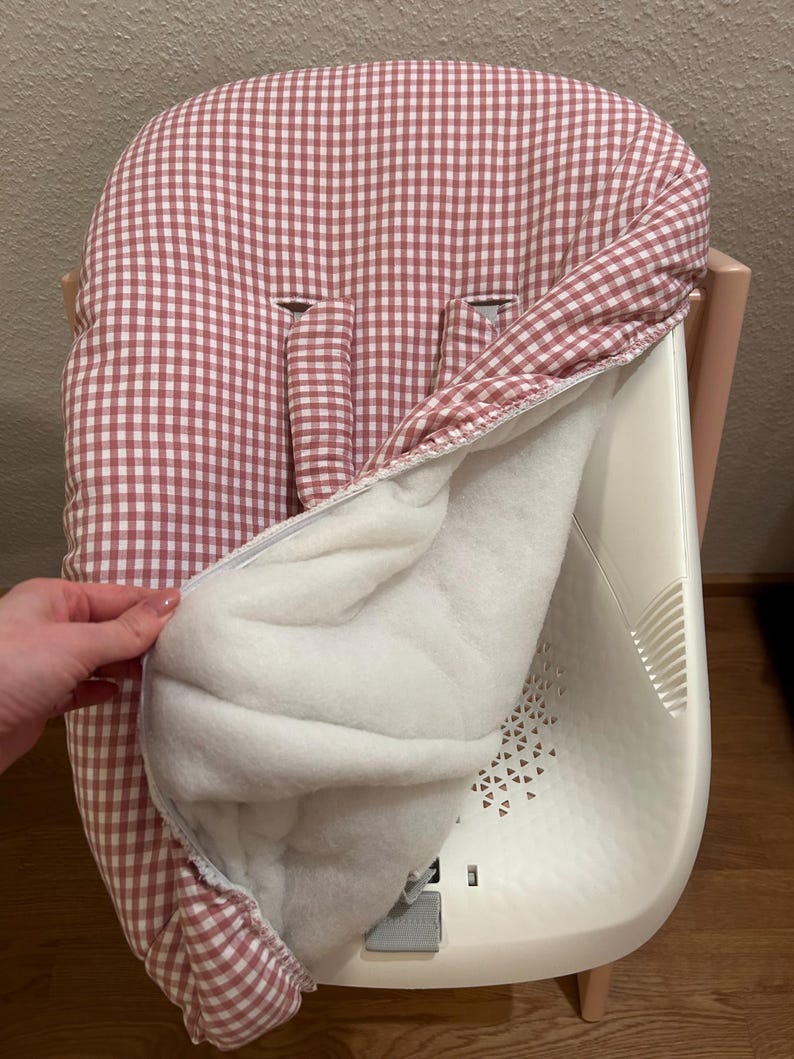 May include: A baby high chair with a removable, padded seat cover. The cover features a red and white gingham pattern on the exterior and a soft white interior. The high chair is white and has a light wood-colored base.