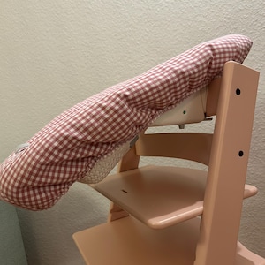 May include: A light pink wooden high chair with a red and white checkered cover. The high chair has a seat and footrest. The cover is made of a soft fabric and is designed to fit over the chair's back and headrest.