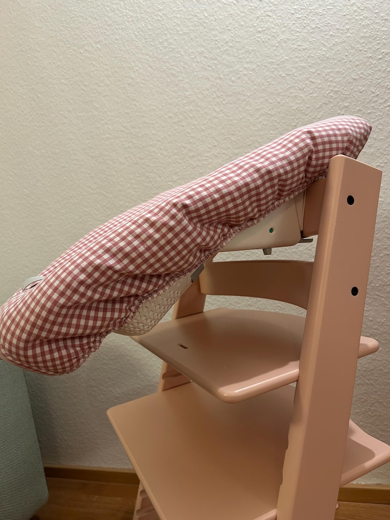 May include: A light pink wooden high chair with a red and white checkered cover. The high chair has a seat and footrest. The cover is made of a soft fabric and is designed to fit over the chair's back and headrest.