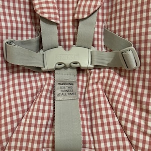 May include: Close-up of a baby high chair harness with a red and white gingham pattern. The gray harness has a plastic buckle with the brand name "Stokke" and a warning label that reads "WARNING: USE THIS HARNESS AT ALL TIMES."