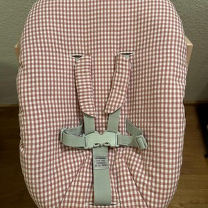 May include: A baby high chair seat cushion with a red and white gingham pattern. The seat has a five-point harness with a gray buckle and straps. The text "WARNING USE THIS HARNESS AT ALL TIMES" is visible.