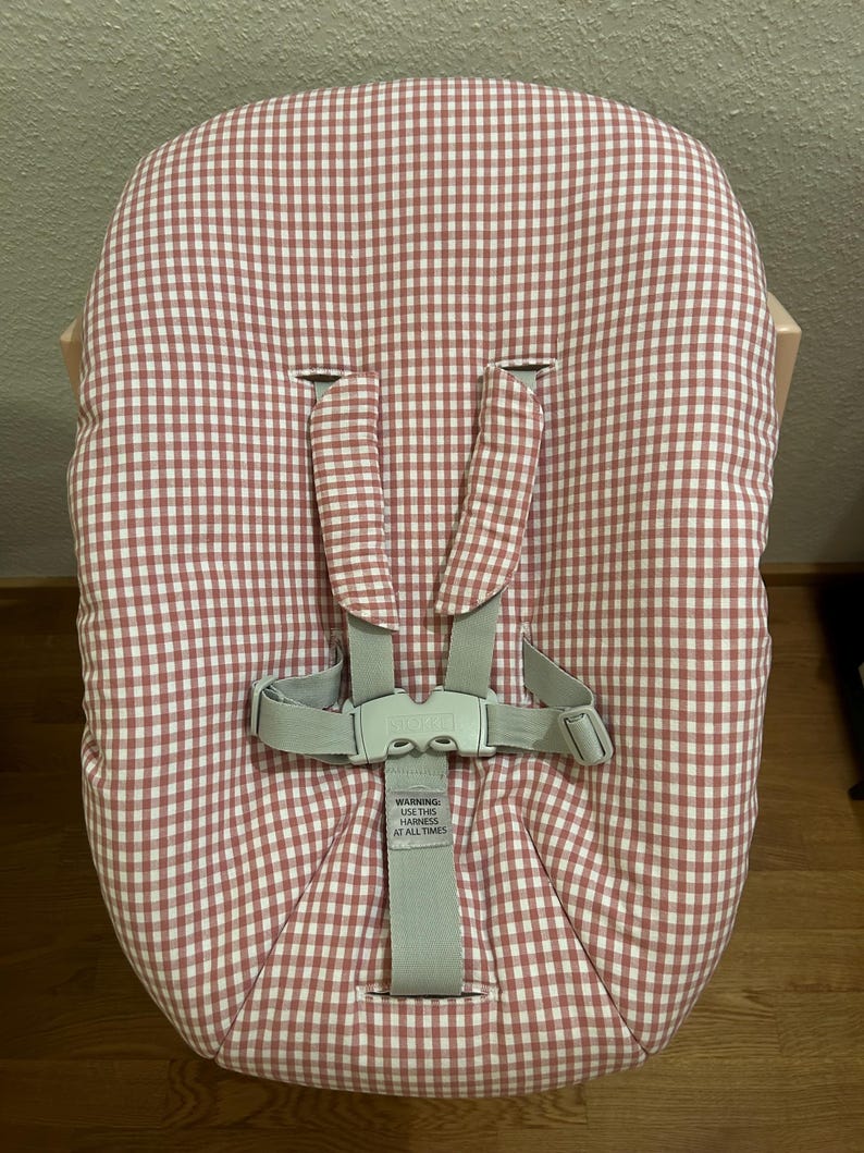 May include: A baby high chair seat cushion with a red and white gingham pattern. The seat has a five-point harness with a gray buckle and straps. The text "WARNING USE THIS HARNESS AT ALL TIMES" is visible.