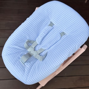 May include: A light pink high chair with a blue and white gingham patterned seat cover. The seat cover has gray straps and a buckle for safety. The high chair is made of plastic and is designed for babies and toddlers.