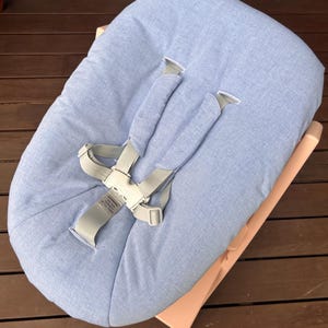 May include: A baby bouncer seat featuring a light blue fabric cover with a textured weave. The seat includes a light gray harness system with padded shoulder straps and a central buckle. A small warning label is visible on the center strap.