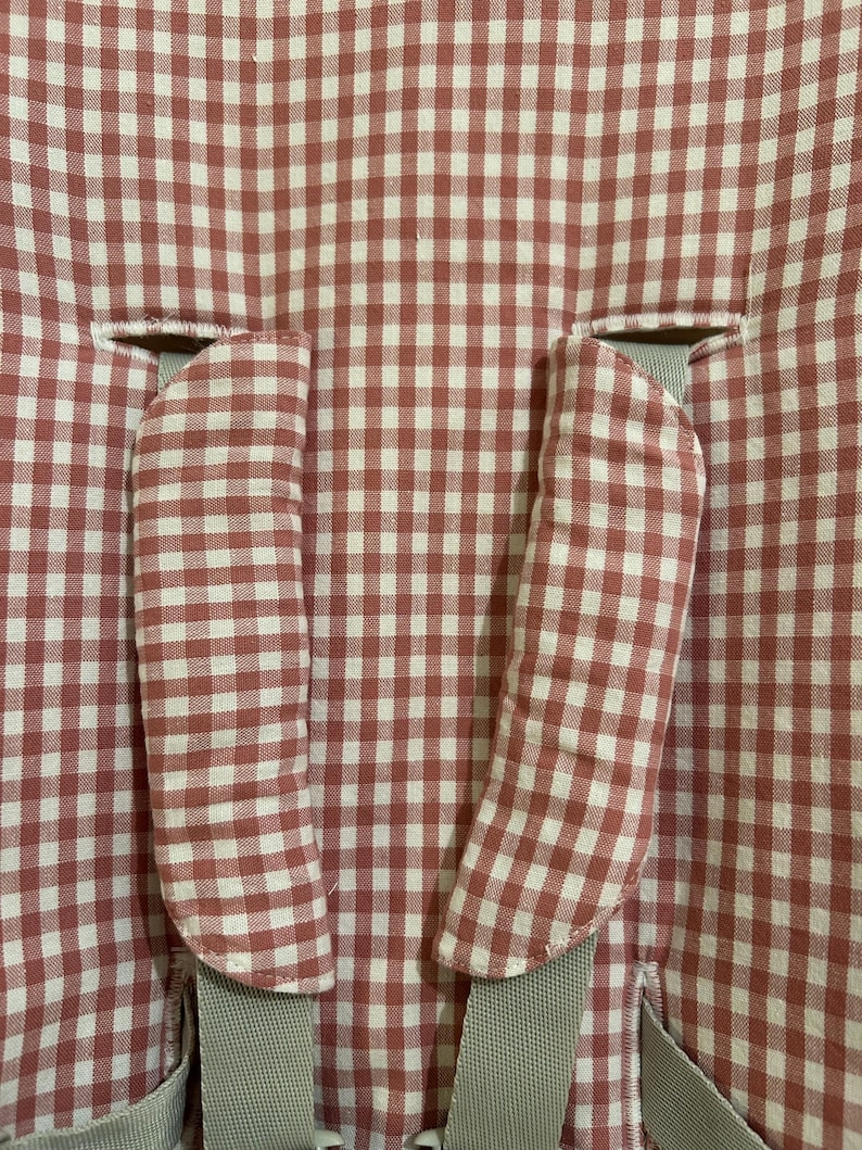 May include: Close-up of a baby high chair seat cover. The cover features a red and white gingham pattern. Two padded armrest covers are visible, along with gray straps for securing the child. The fabric is a soft, woven material.