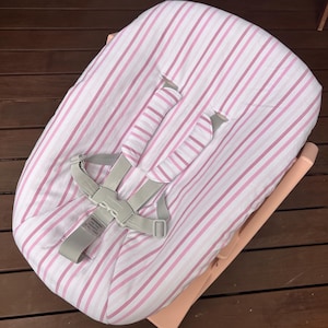 May include: A baby bouncer seat featuring a white fabric cover with vertical pink stripes. The seat includes a light gray five-point safety harness with padded shoulder straps and a central buckle. The frame is a light peach color with a curved base.