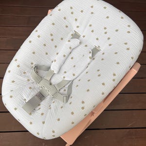 May include: A baby bouncer featuring a white fabric cover with a repeating pattern of small tan four-leaf clovers. The bouncer includes a light gray safety harness with padded shoulder straps and a central buckle with the text WARNING USE THE HARNESS AT ALL TIMES. The frame is a solid light pink color.