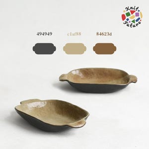 Può includere: Due piatti ovali in ceramica con piccole maniglie laterali. Le superfici interne presentano una smaltatura maculata beige e marrone, mentre le basi esterne sono grigio antracite scuro. L'immagine include i codici colore 494949, c1af88, 84623d e il testo Knit Future.