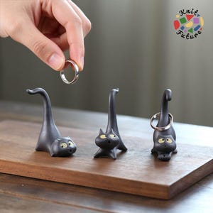 Puede incluir: Tres figuras de gato de cerámica negra mate diseñadas como soportes para anillos, cada una con una cola alargada para sostener joyas. Los gatos tienen ojos amarillos grandes y sorprendidos y bocas pequeñas. Un anillo de plata es sostenido por una cola.