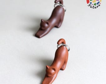 May include: Two cat-shaped ceramic figures in dark brown and reddish-brown, each featuring a silver-coloured metal ring positioned around the tail. The figures are in a stretching pose with heads lowered. The logo Knit Future is visible in the corner.