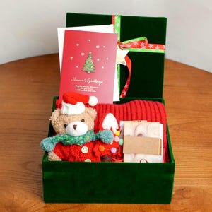Puede incluir: Una caja de regalo de terciopelo verde atada con una cinta roja y verde, que contiene una bufanda roja de punto, un oso de peluche con gorro y bufanda de Papá Noel y una tarjeta de Navidad roja con el texto "Season's Greetings".