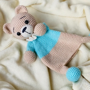 Pode incluir: Um cobertor de segurança de urso de pelúcia de crochê feito à mão. O urso tem uma cabeça bege com olhos pretos e um nariz costurado em preto. O cobertor é azul claro e bege com detalhes de bolas azuis. O brinquedo de pelúcia está sobre um cobertor creme.