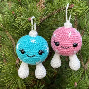 May include: Two handmade crochet Christmas ornaments. One is light blue, the other is pink, both with white snowflake accents and smiling faces. They hang from white strings against a backdrop of green pine needles.