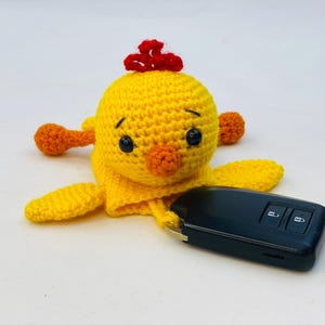 May include: A handmade yellow crochet chick with orange feet, beak, and a red crest. The chick is lying down next to a black car key fob. The chick has black eyes.