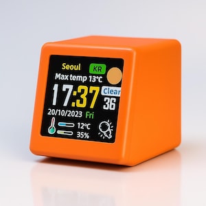 Könnte beinhalten: Eine orangefarbene, würfelförmige Digitaluhr, die die Uhrzeit 17:37 und das Datum 20.10.2023 anzeigt. Der Bildschirm zeigt Wetterinformationen für Seoul, einschließlich einer Höchsttemperatur von 13°C. Die Uhr hat eine klare Anzeige.