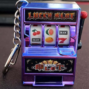 Può includere: Portachiavi con mini slot machine con finitura metallica viola e blu. Il portachiavi presenta il testo "LUCKY SLOT" e "CASINO SLOT". I rulli mostrano ciliegie, un'anguria, un'arancia e un sette fortunato.