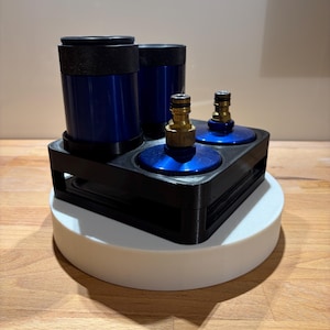 Op de afbeelding: Een zwarte kunststof houder met twee hoge blauwe cilindrische containers en twee kortere blauwe ronde basissen, elk voorzien van een messingkleurige metalen snelkoppeling voor slangen. De onderdelen zijn geplaatst in een vierkant frame.