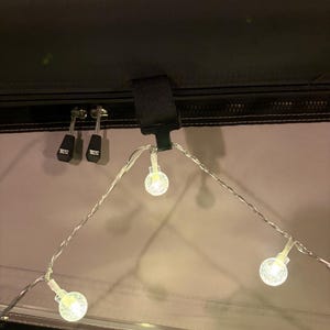 Op de afbeelding: Een lichtsnoer met heldere, bolvormige LED-lampjes met een textuur van kleine belletjes aan de binnenzijde. De lampjes hangen aan een zwarte plastic clip bevestigd aan een donkere stoffen ondergrond met twee ritstrekkers voorzien van de tekst TELTA.