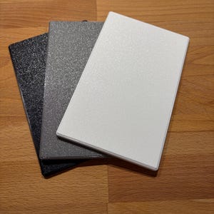 Könnte beinhalten: Drei rechteckige Platten mit einer texturierten, körnigen Oberfläche. Die Platten sind in einem überlappenden Stapel angeordnet und bestehen aus einer schwarzen Platte, einer mittelgrauen Platte und einer hellweißen Platte oben.