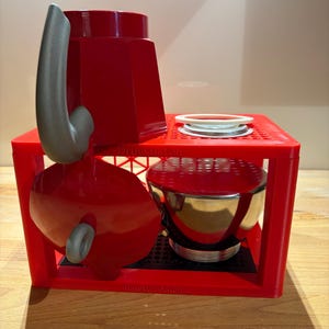 Può includere: Un supporto in plastica rossa a forma di cassetta con la scritta Druckkasten e Bialetti Edition. Contiene una caffettiera espresso rossa con manico grigio e base in metallo argentato. Un anello bianco poggia sulla superficie del supporto.