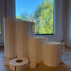 Op de afbeelding: Een set van vier witte cilindrische houders van verschillende hoogtes, elk voorzien van een handvat aan de zijkant en de tekst "by Druckhafen" in reliëf. Een ronde witte deksel met een geperforeerde bovenkant en twee losse clips zijn inbegrepen.