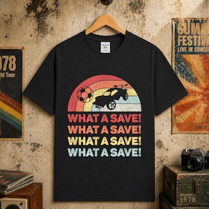 May include: Black t-shirt with a retro design featuring a soccer ball, a car, and a rainbow. The text "WHAT A SAVE!" is repeated four times in a colorful font. The shirt is hanging on a wooden hanger.