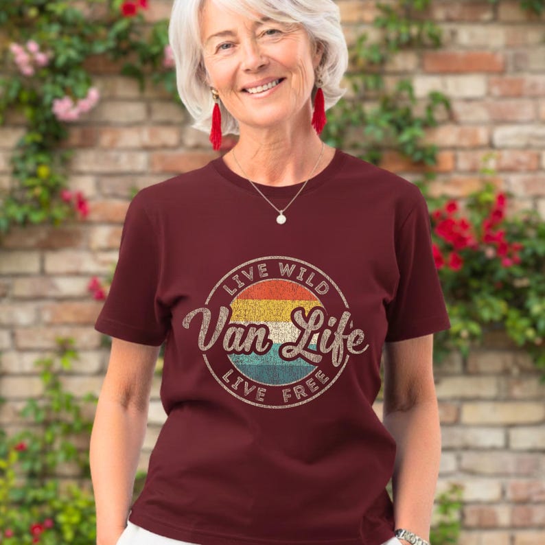 May include: A maroon t-shirt with a circular graphic that reads "Van Life" in a vintage style. The graphic features a retro color scheme of yellow, orange, blue, and white. The text "Live Wild, Live Free" surrounds the main text.
