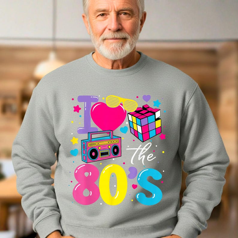 Op de afbeelding: Een grijze sweatshirt met een kleurrijk jaren 80-thema. Het ontwerp bevat een roze hart, een boombox, een Rubik's Cube en de tekst "I love the 80's" in levendige kleuren. Een casual kledingstuk.